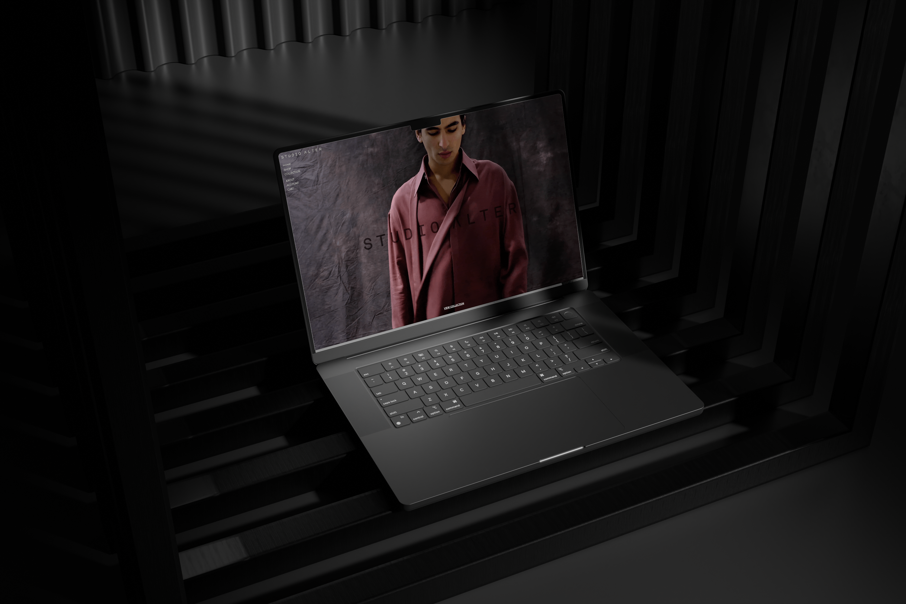 A dark gray open laptop placed on a black slatted surface, with a Studio Alter homepage showing a person in a red jacket. The product itself appears to be the laptop, photographed in a moody dark studio setting.