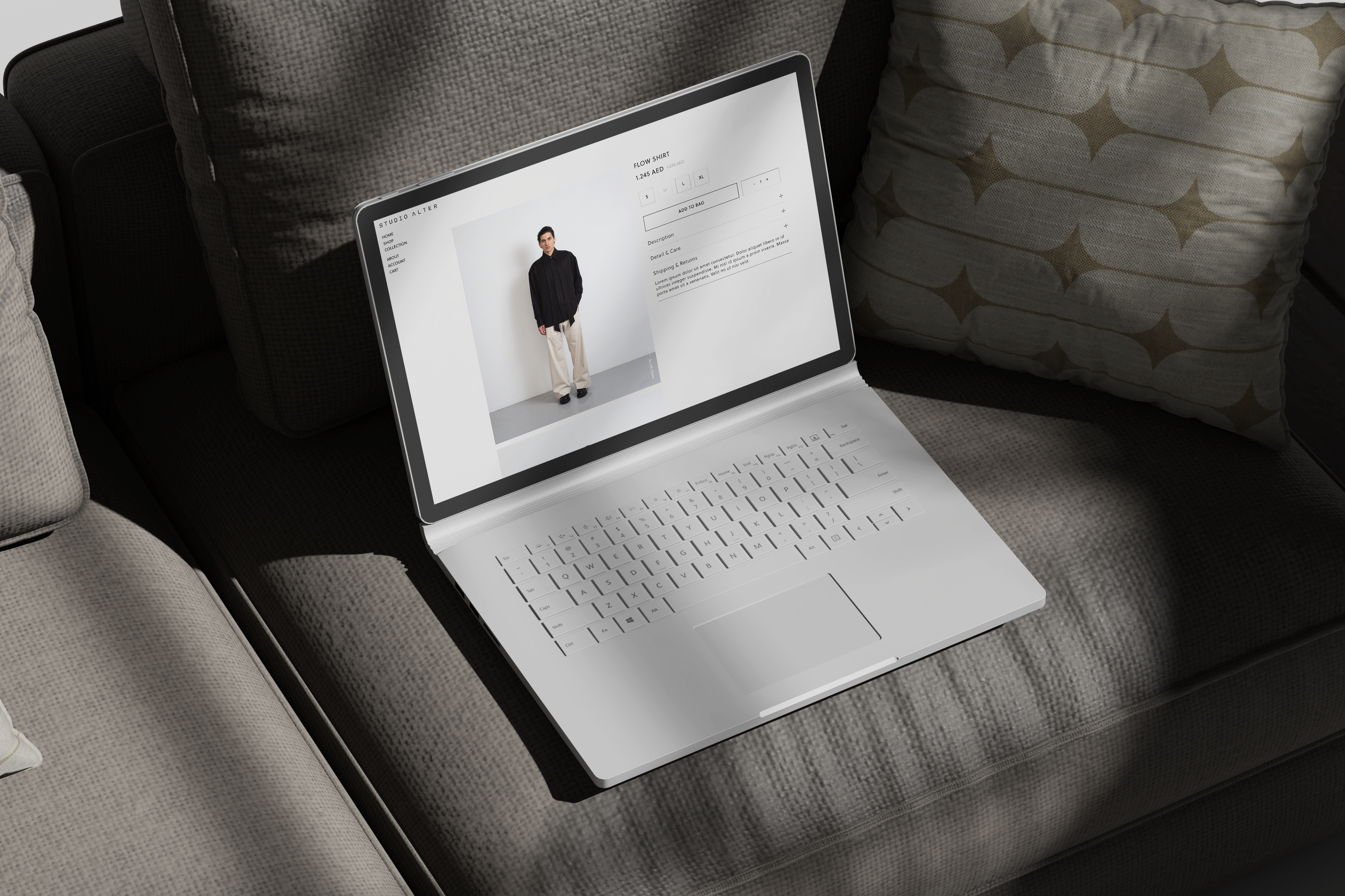 A silver laptop sits open on a gray upholstered sofa, displaying a Studio Alter fashion e-commerce webpage with a model photo. The scene is lit by natural light with soft shadows, and a patterned throw pillow is visible in the background.